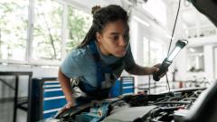 A little care for your vehicle. Young african american woman, professional female mechanic looking, examining under hood of car with torch at auto repair shop